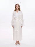 A woman modeling a long white silk-like robe with a tie waist and lace-trimmed cuffs, standing against a plain white studio background.