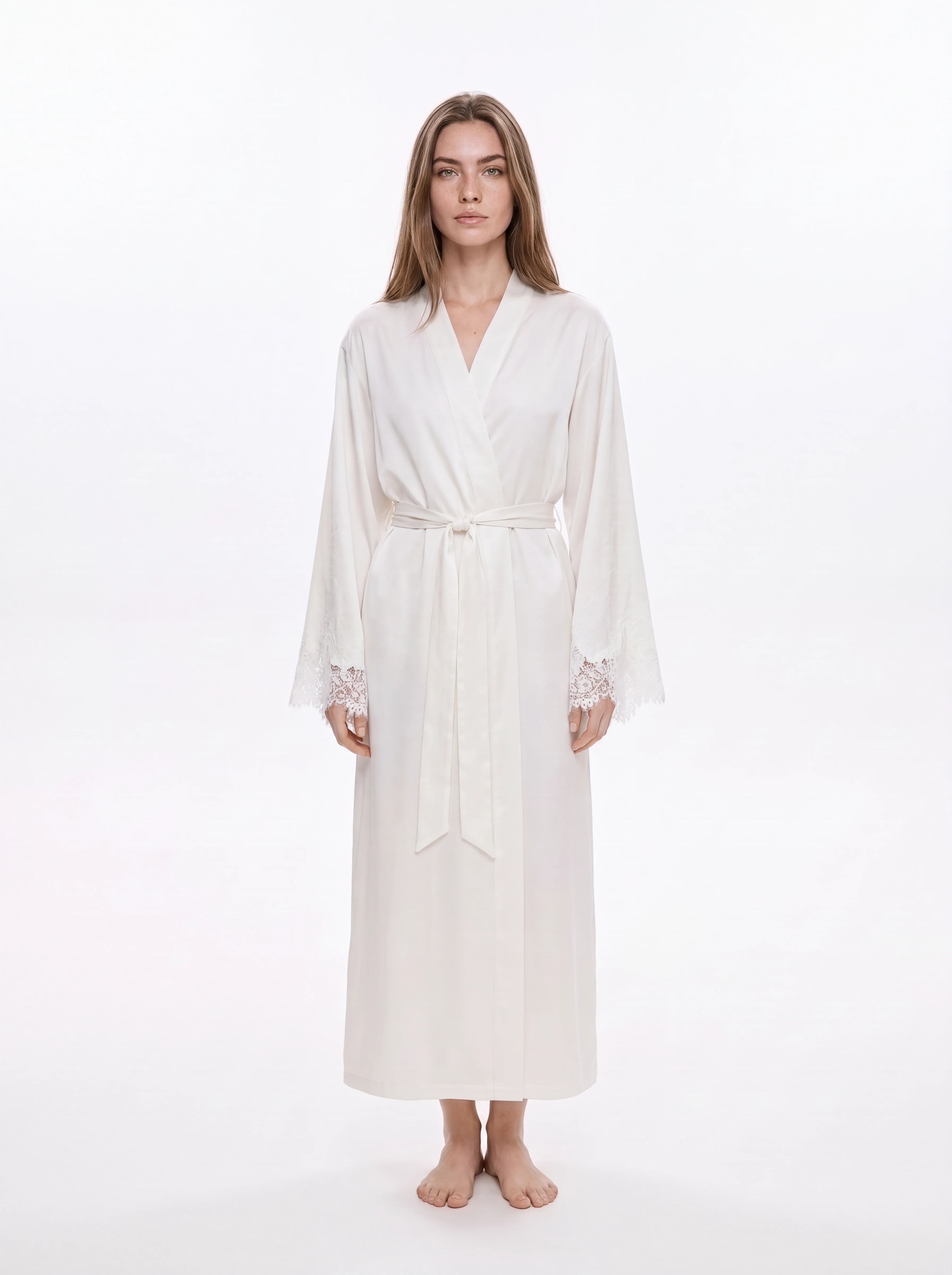 A woman modeling a long white silk-like robe with a tie waist and lace-trimmed cuffs, standing against a plain white studio background.