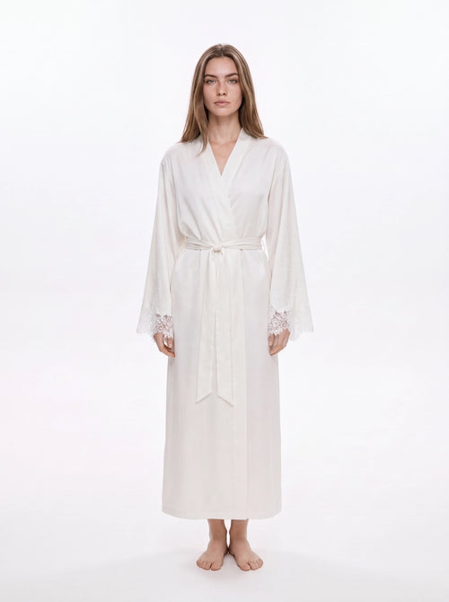 A woman modeling a long white silk-like robe with a tie waist and lace-trimmed cuffs, standing against a plain white studio background.