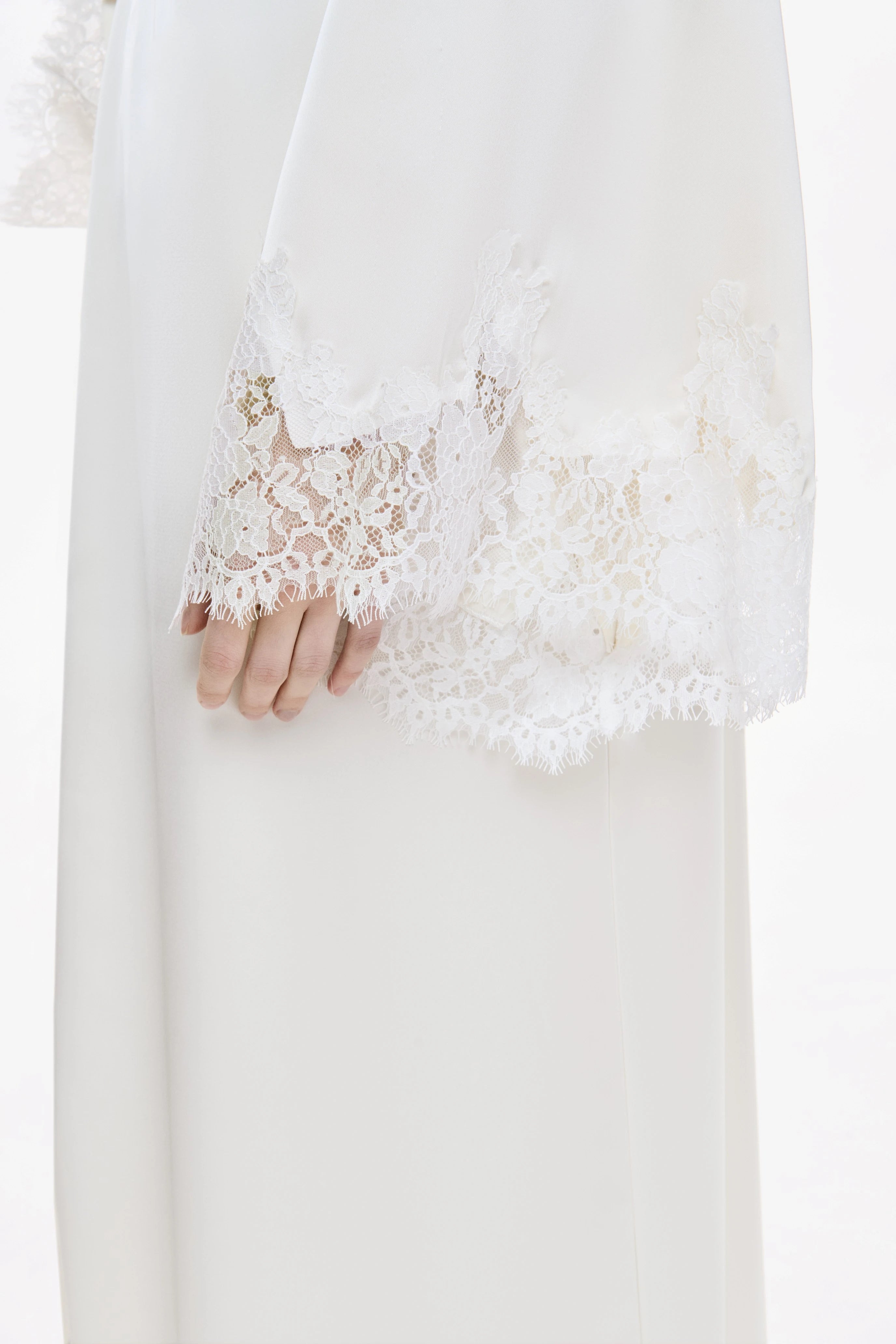 A close-up of a white flowing garment with delicate lace trim and a hand resting near the hem, shown against a very light neutral background.