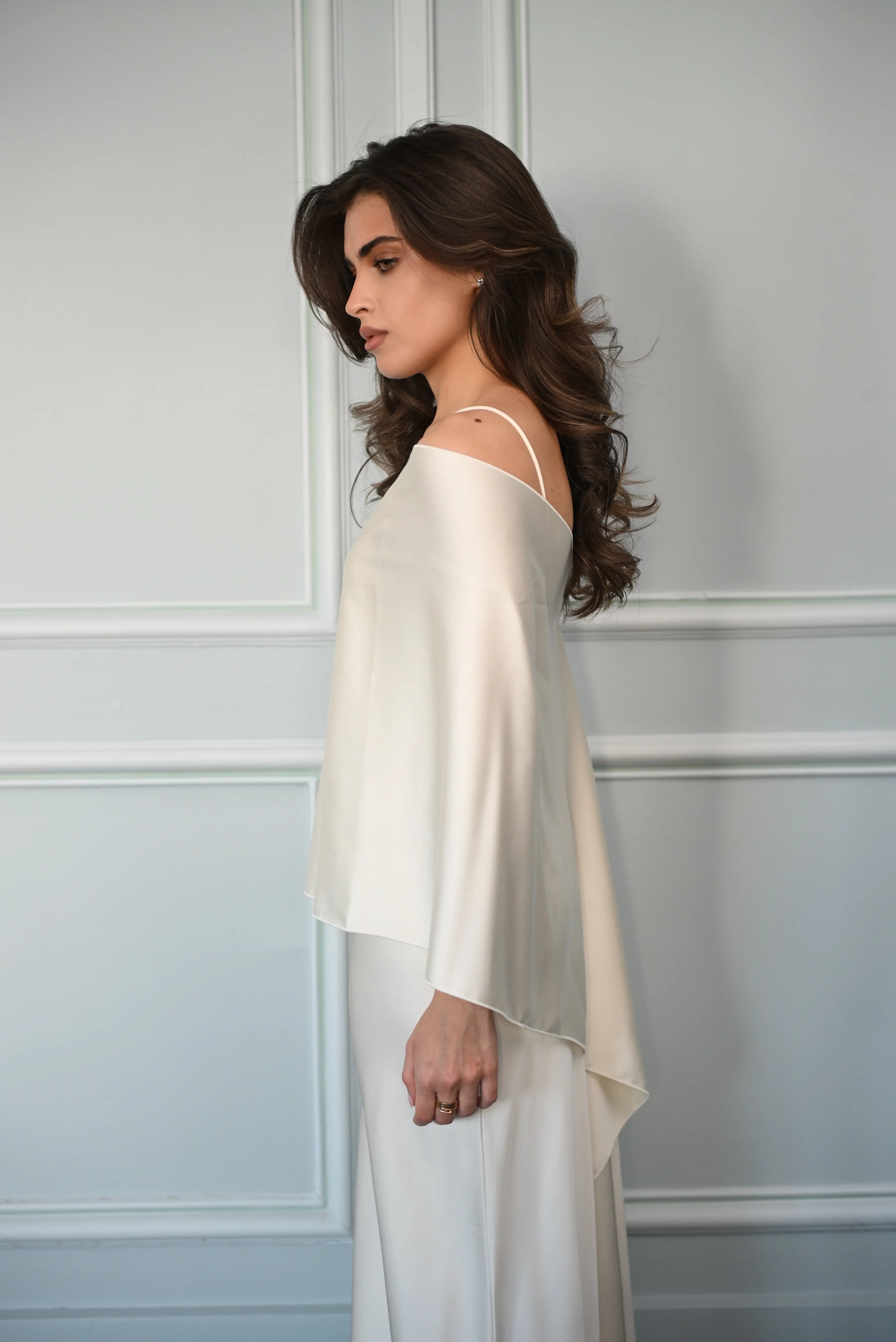 Woman wearing a white off-shoulder top and flowing pants against a light gray wall.