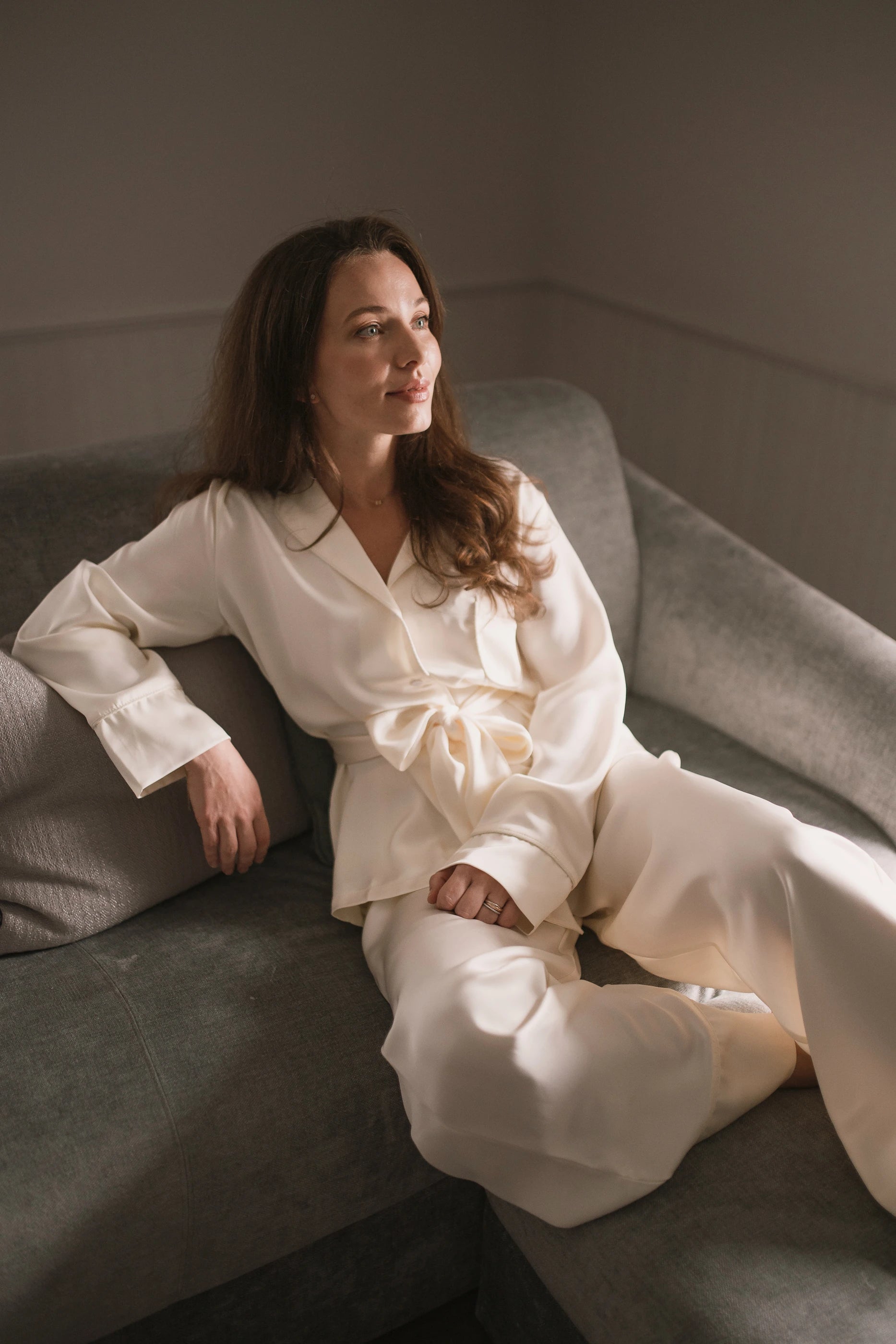 Woman in a ivory white pyjama sitting on a couch in a softly lit room.