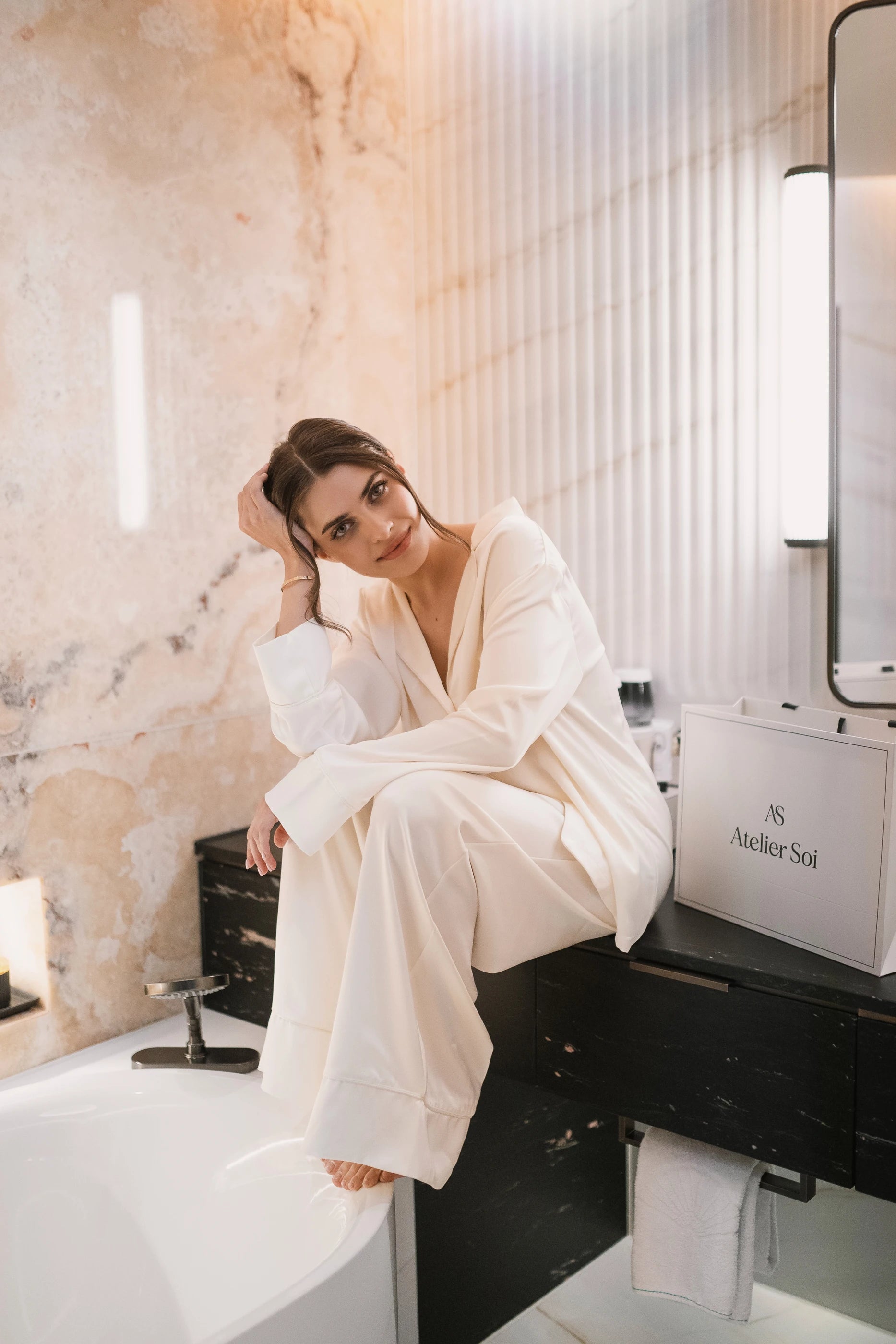 Woman in a white pyjama sitting on a black bench in a bathroom with marble walls.