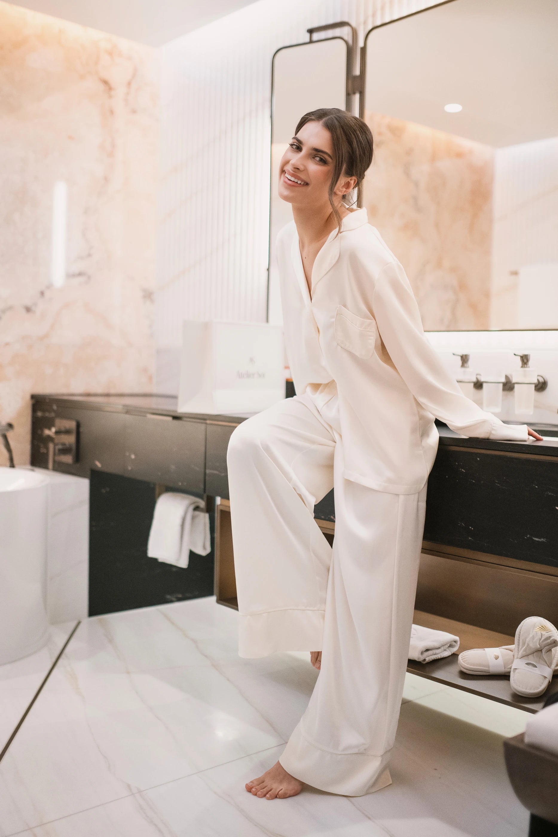 Woman in a ivory white satin pyjama sitting in a luxurious bathroom.