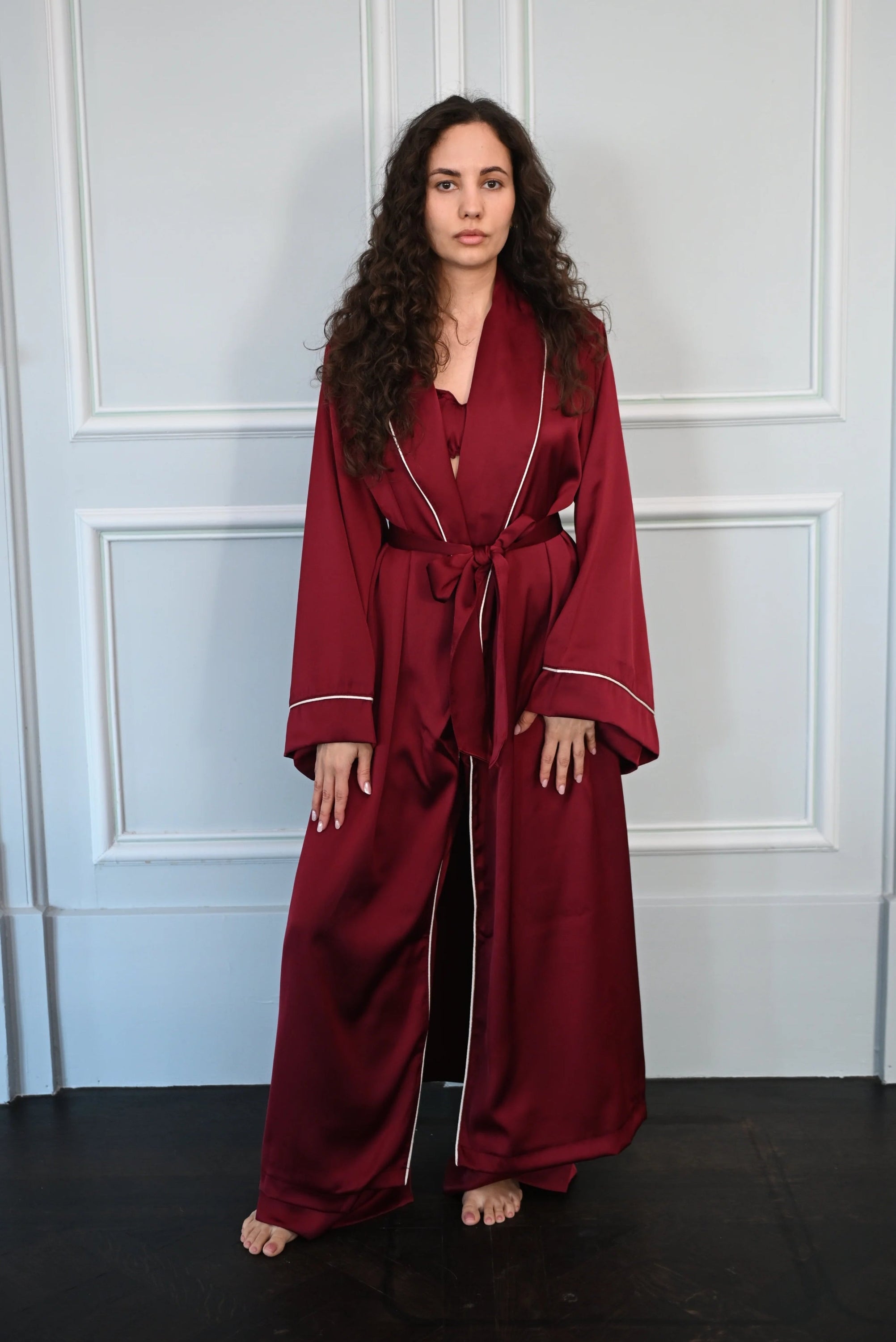 Woman wearing a burgundy robe standing against a white paneled wall.