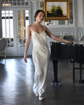 Woman in a white dress standing next to a black piano in an elegant room with chandeliers and a painting.