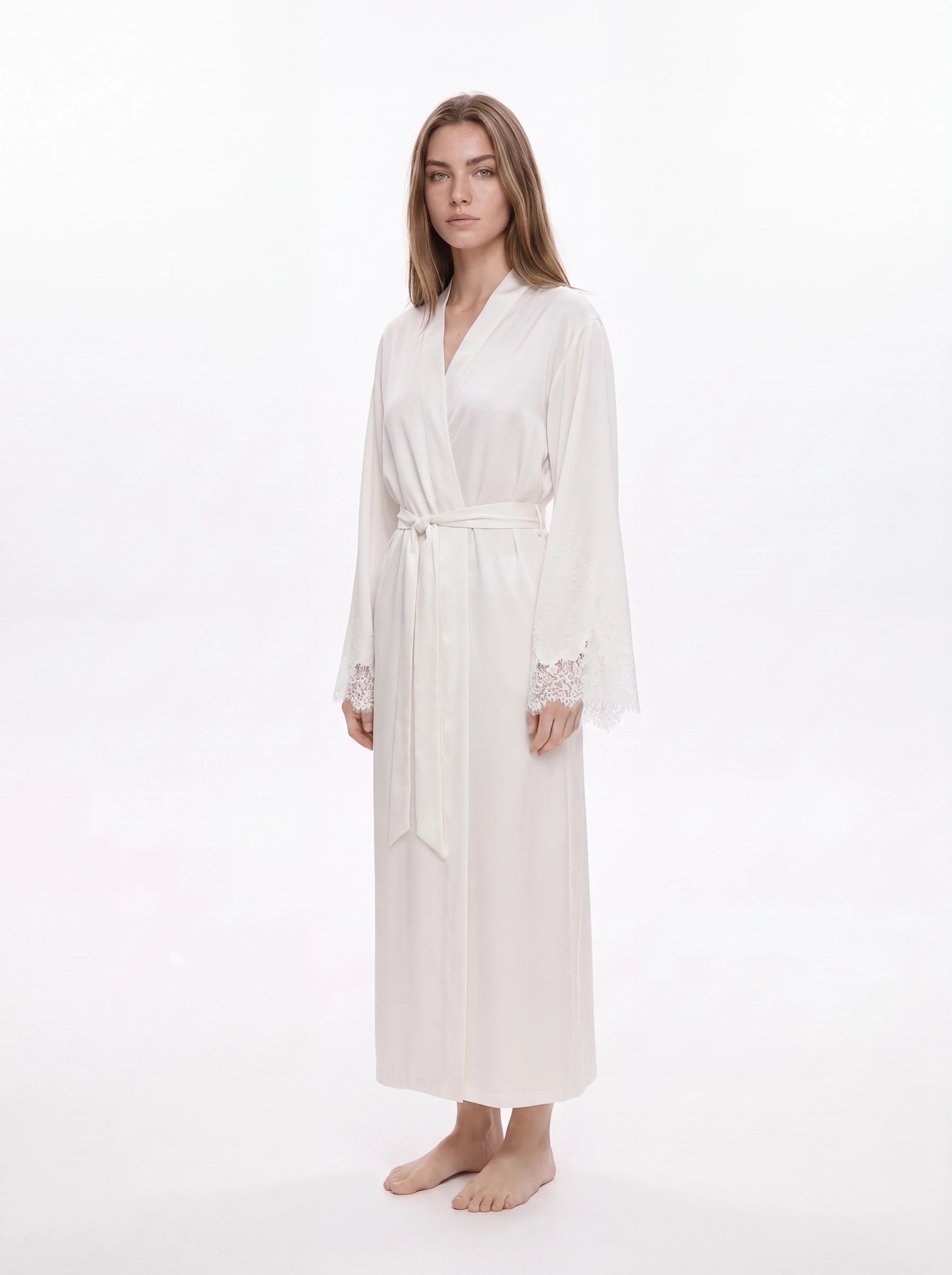 A woman modeling a long white silk-like robe with a tie waist and lace cuffs, standing against a plain white studio background.