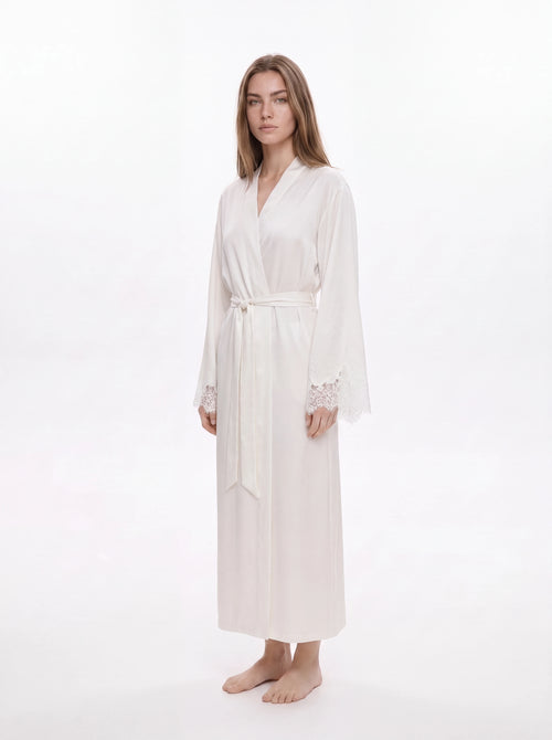 A woman modeling a long white silk-like robe with a tie waist and lace cuffs, standing against a plain white studio background.