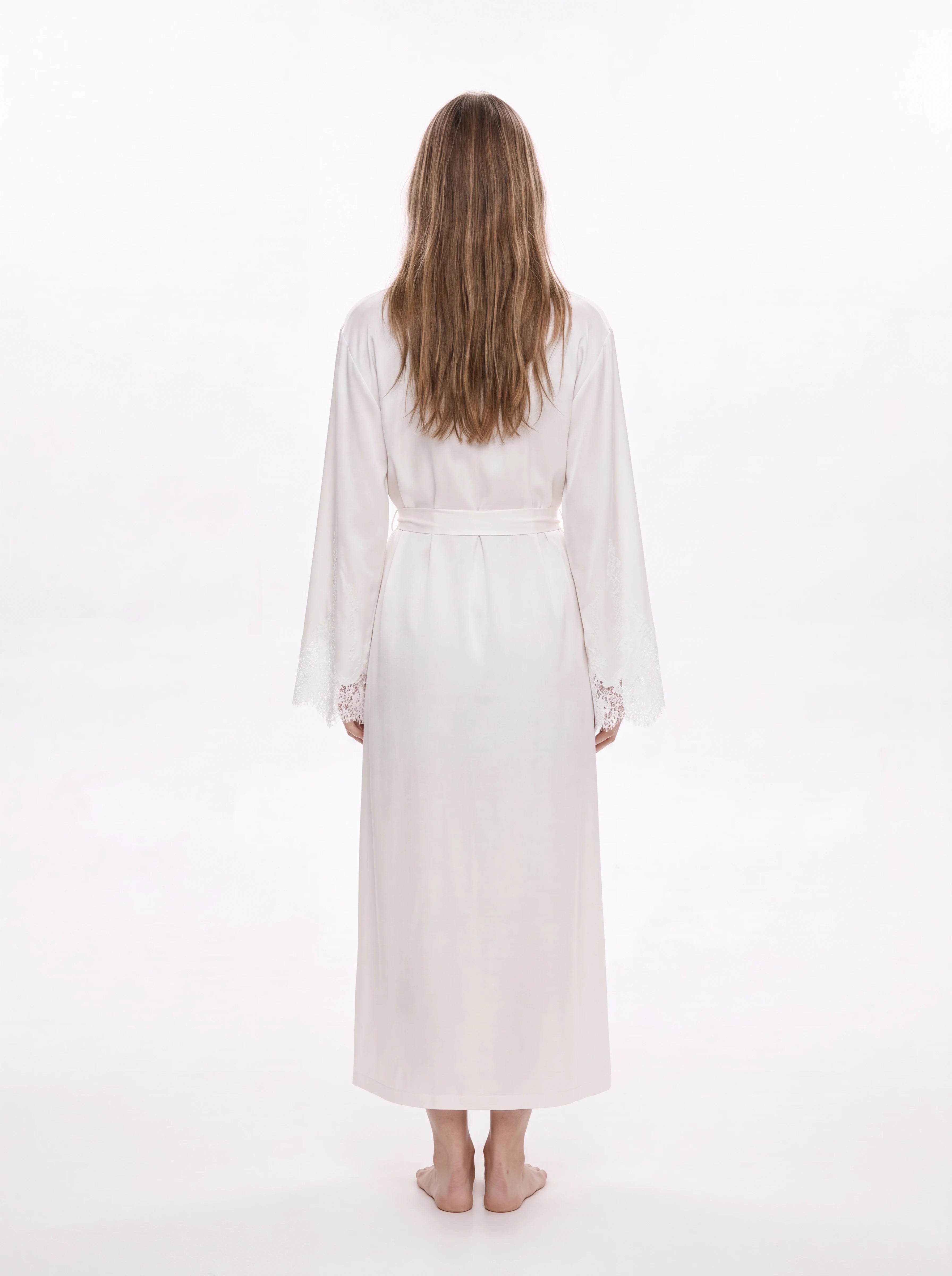 A woman viewed from behind wearing a long white robe with lace cuffs, standing against a plain white studio background.